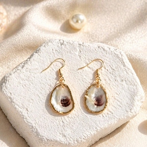 May include: A pair of seashell earrings with gold-colored trim and ear hooks. The earrings are oval-shaped, with a white and purple interior. The earrings are displayed on a white, textured surface, with pearls and a seashell in the background.