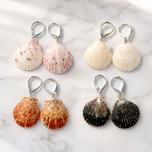 Puede incluir: Cuatro pares de pendientes de concha con cierres de palanca plateados. Las conchas varían en color, incluyendo rosa, crema, naranja y negro, cada una con una apariencia natural y texturizada. Los pendientes se muestran sobre una superficie blanca.