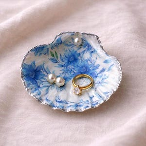 Op de afbeelding: Een schelpvormige sieradenschaal met een blauw bloemmotief en zilveren rand. De schaal bevat een gouden ring met een heldere steen en twee paar witte parels. De schaal ligt op een crèmekleurige stof.