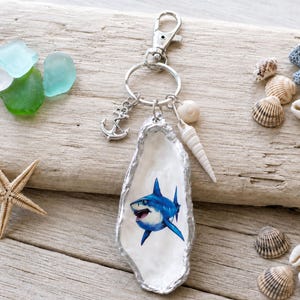 Op de afbeelding: Een sleutelhanger met een zilverkleurige karabijnhaak en ring, versierd met een kleine zilveren ankerbedel, een witte spiraalschelp en een oesterschelp beschilderd met een blauwe haai. Omringd door zeeglas en diverse schelpen.