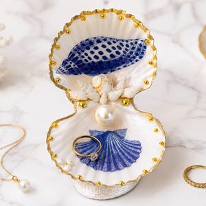 May include: A decorative open seashell trinket dish featuring gold-painted edges and small gold beads along the rim. The interior displays blue shell illustrations, a central white pearl, and smaller white shells. A gold ring rests inside.