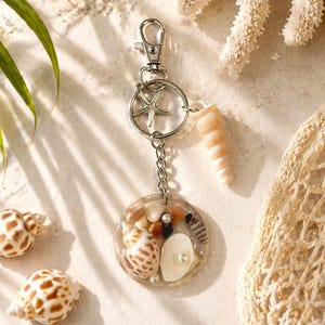 以下が含まれることがあります： 透明な樹脂のチャームが付いたシルバーのキーホルダー。チャームには貝殻、真珠、小さな石が詰まっています。キーホルダーにはヒトデのチャームと小さな貝殻のペンダントが付いています。砂浜の表面には、さらに貝殻が散らばっています。