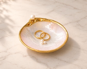 Puede incluir: Un plato para joyas con forma de concha, interior blanco nacarado y borde dorado texturizado. Una perla blanca está fijada en el borde, mientras que dos anillos dorados y dos pequeñas perlas blancas sueltas descansan en el interior.