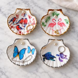 May include: Four white scallop shell trinket dishes with gold-painted edges and a single pearl accent. Each dish features unique colorful illustrations: red birds on branches, pink flamingos with green leaves, blue butterflies, and sea turtles.