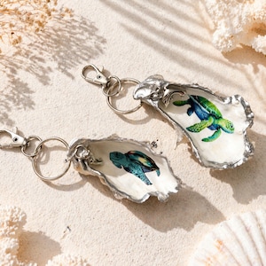 May include: Two keyrings with sea turtle designs inside oyster shells. The shells have a silver-coloured metal trim and are attached to a silver-coloured metal clasp and ring. The turtles are blue and green.