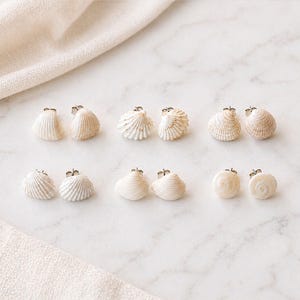 Puede incluir: Un conjunto de pendientes de concha en varias formas y texturas. Los pendientes son blancos y beige, dispuestos sobre una superficie de mármol con una tela de color neutro en el fondo. Los pendientes son pequeños y delicados.