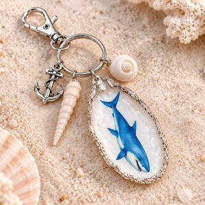May include: A silver-toned keychain featuring a metal lobster clasp and ring. Attached are a small silver anchor charm, a long spiral seashell, a round white seashell, and an oval pendant with a blue shark illustration on a white background.