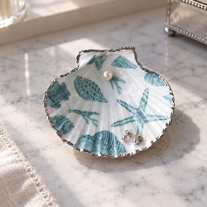 May include: A white scallop shell trinket dish featuring a teal blue pattern of starfish, fish, and coral motifs. The shell has a metallic silver-toned rim and contains a single white pearl and a pair of small silver stud earrings inside.