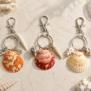 May include: Three silver-toned metal keychains, each featuring a circular ring and a lobster clasp. The keychains are adorned with natural seashells in shades of orange, red, and cream, along with small silver-toned metal anchor charms.