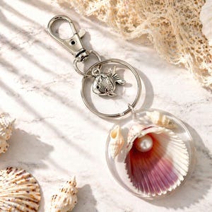 May include: A silver keyring with a fish charm and a clear, round pendant. The pendant features a seashell with a pearl and is surrounded by smaller shells. The background includes more seashells.