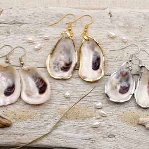 Könnte beinhalten: Drei Paar handgefertigte Ohrringe aus Austernschalen. Zwei Paare haben goldfarbene Verzierungen, ein Paar hat silberfarbene Verzierungen. Die Muscheln weisen natürliche Farbvariationen in Weiß, Braun und Lila auf. Kleine Perlen und Sand sind verstreut.