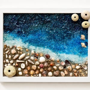 Puede incluir: Una pieza de arte mural rectangular en un marco blanco que presenta una escena de playa creada con vidrio azul triturado para representar las olas del mar y material color arena cubierto con varias conchas pequeñas, erizos y estrellas de mar.