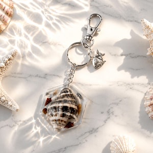 May include: A keychain featuring a clear, hexagonal resin pendant with a seashell and small shell fragments. The keychain has a silver-tone fish charm and a lobster clasp. Seashells and a starfish are arranged around the keychain on a marble surface.