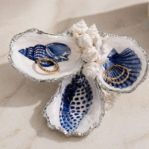 May include: A decorative oyster shell jewelry dish with three sections. Each section is white with a blue seashell design. Two gold rings rest in the dish. Small white seashells are clustered in the center.