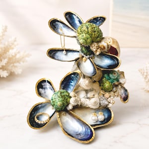 Könnte beinhalten: Ein dekorativer Schmuckhalter aus irisierenden blauen Miesmuschelschalen und grünen Schneckenhäusern, die in Blumenform angeordnet sind. Die Ränder der Schalen sind goldfarben abgesetzt und die Struktur ist mit kleinen weißen Muscheln und Perlen verziert.