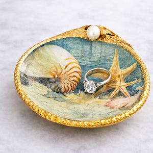 Puede incluir: Un plato decorativo de concha marina con una escena de playa pintada. El plato contiene un anillo de plata con una gema grande y transparente y una perla. La concha tiene un borde dorado y una concha de nautilus y una estrella de mar pintadas.