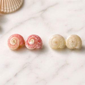 May include: Close-up of four spiral-shaped seashells in shades of pink and cream. Two are a vibrant pink, and two are a creamy white. The shells are arranged on a white marble surface, suggesting a coastal theme.