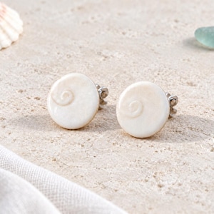 May include: A pair of round stud earrings featuring a smooth, off-white shell surface with a subtle, natural spiral pattern embossed on each piece. The earrings are secured with metal posts and backings, showcasing a clean, circular design.