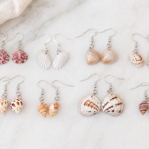 May include: A collection of seashell earrings with silver-colored hooks. The earrings feature various shell shapes and colors, including pink, white, tan, and brown. The earrings are displayed on a white marble surface.