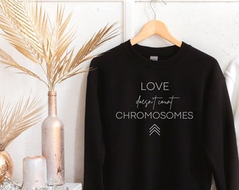 May include: Two sweatshirts, one black and one white, with the text "LOVE doesn't count CHROMOSOMES" printed on the front. The sweatshirts are hanging on hangers against a white wall.