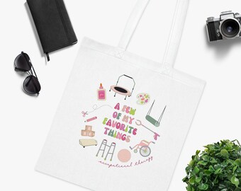 Op de afbeelding: Witte tote bag met een roze en groen ontwerp met verschillende objecten, waaronder een trampoline, een schommel, een palet met verf, een bal, een rollator en een rolstoel. De tekst "A few of my favorite things" is geschreven in roze en groene letters. De tekst "occupational therapy" is geschreven in roze letters aan de onderkant van de tas.
