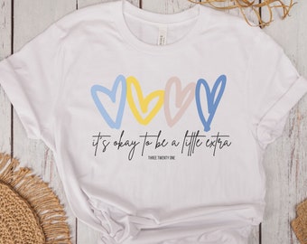 Può includere: Maglietta bianca con una grafica di tre cuori colorati e il testo "it's okay to be a little extra" con il nome del marchio "Three Twenty One" sotto.