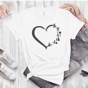 May include: White t-shirt featuring a grey heart design. The heart is filled with various symbols representing occupational therapy, including a wheelchair, a walker, and a person using a walking stick. The text "I ♥ OT" is inside the heart.