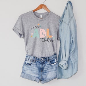 May include: A gray t-shirt with the text "I can't ADL today" in colorful letters with stars. The shirt is on a hanger with a pair of denim shorts and a denim jacket.