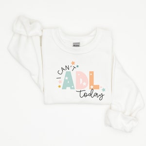 May include: White long-sleeved sweatshirt with the text "I can't ADL today" in colorful letters with stars around the text.