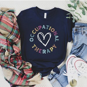 May include: A navy blue t-shirt with a colorful graphic that says "Occupational Therapy" in a circle with a heart in the center.