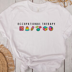 May include: White t-shirt with the text "Occupational Therapy" and a row of colorful icons representing common occupational therapy tools, including a puzzle, stacking rings, pencil, buttons, a block, and a ball.