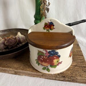 Może przedstawiać: Vintage ceramiczna solniczka z drewnianą pokrywą, zdobiona jabłkami, winogronami i liśćmi. Kremowa solniczka stoi na drewnianej desce. W tle zielony młynek do pieprzu. Akcesorium kuchenne.