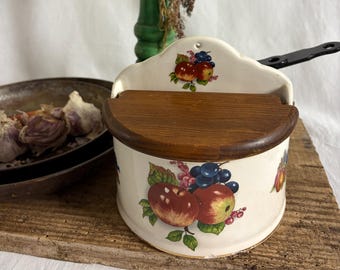 May include: A vintage ceramic salt box with a wooden lid, featuring a hand-painted design of apples, grapes, and leaves. The salt box is cream-colored and sits on a wooden board. A green pepper grinder is in the background.