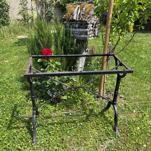 Peut inclure: Structure de table en fer forgé noir avec un plateau rectangulaire et des pieds ornés. Le cadre est installé dans un espace extérieur herbeux avec un nichoir et un rosier rouge en arrière-plan.
