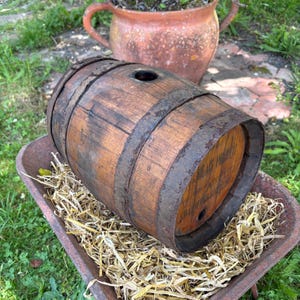 Peut inclure: Un petit tonneau en bois vieilli avec des cerceaux métalliques. Le tonneau est brun foncé et percé d'un trou sur le dessus. Il repose dans une brouette rouillée remplie de paille, avec un pot en terre cuite en arrière-plan.