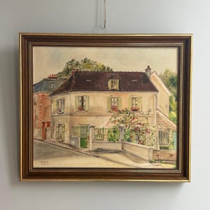 Peut inclure: Peinture à l'aquarelle encadrée d'une scène de village européen. L'œuvre représente un bâtiment à deux étages avec un toit brun, des volets verts et un petit jardin. La peinture est encadrée dans un cadre en bois brun foncé.