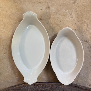 Peut inclure: Deux plats de cuisson ovales en céramique blanc cassé avec une finition lisse et brillante. Chaque plat présente une poignée décorative en forme de coquille aux deux extrémités. Le plus grand plat est à gauche, le plus petit à droite.