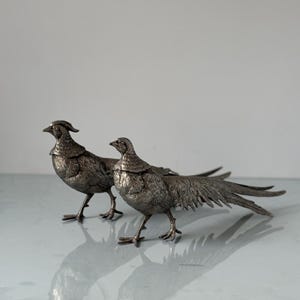 Peut inclure: Deux figurines de faisans argentés détaillés. Les oiseaux se tiennent sur une surface réfléchissante, avec leurs longues plumes de queue déployées. Les figurines présentent des détails de plumes complexes et sont orientées dans la même direction.