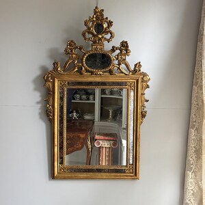 Puede incluir: Un espejo de pared rectangular con un marco de color dorado con intrincados detalles tallados, una cresta decorativa en la parte superior y un inserto de espejo ovalado más pequeño sobre el panel de vidrio principal, reflejando el interior.