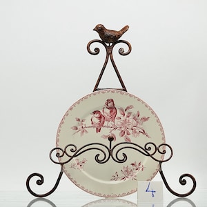 May include: A decorative plate on a bronze metal stand. The plate has a red and white floral and bird design. The stand features a bird and decorative scrollwork. The plate is approximately 25 cm in diameter.