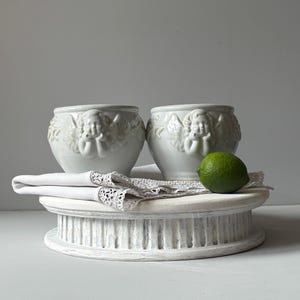 Peut inclure: Deux bols en céramique blanche ornés de figurines de chérubins en relief avec des ailes sur l'extérieur. Un tissu blanc plié avec une bordure en dentelle est placé sous les bols, et un citron vert repose sur le tissu à côté des récipients.