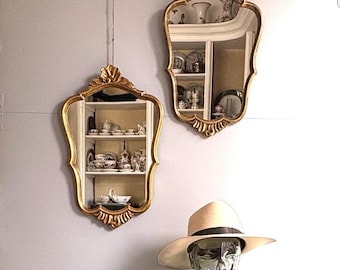 May include: Two gold-toned wall mirrors with ornate, scalloped frames featuring shell-like carvings at the top and bottom. A beige wide-brimmed hat rests on a clear glass mannequin head, positioned below the mirrors on a dark green surface.