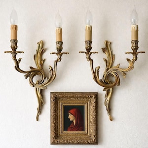 Puede incluir: Dos apliques dorados ornamentados, cada uno con tres bombillas en forma de vela, montados en una pared blanca. Debajo, un retrato enmarcado de una persona con un tocado rojo.