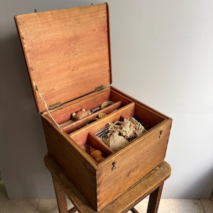 Peut inclure: Une boîte à couture rectangulaire en bois avec un couvercle à charnière, comprenant plusieurs compartiments intérieurs. La boîte contient divers accessoires de couture, notamment des bobines de fil, de la dentelle et de petits outils.