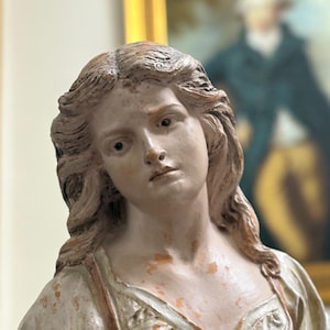 Puede incluir: Busto detallado de una mujer con cabello largo y ondulado y una expresión serena. La escultura está pintada en tonos apagados de blanco, beige y marrón, con signos visibles de edad y desgaste. La mujer lleva un vestido con escote.