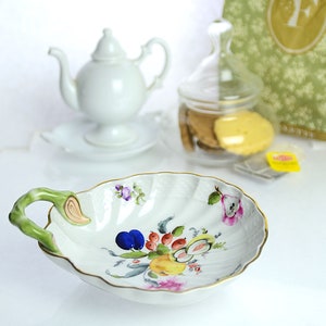 May include: A white porcelain serving dish with a scalloped edge and a green handle. The dish is decorated with a colorful floral and fruit design, including plums, pears, and berries. A white teapot and a glass jar with cookies are in the background.