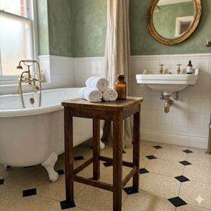 May include: A square wooden stool with four legs stands in a bathroom. On top of the stool are three rolled white towels stacked in a pyramid shape and a small amber glass bottle with a cork stopper. The stool has a dark brown wood finish.