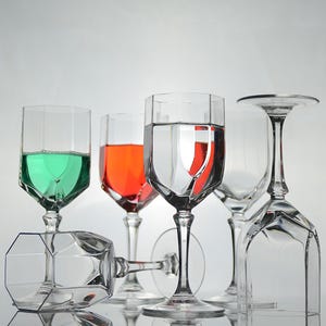 May include: Five clear glass wine glasses, two with green and red liquid, one with water, and two empty. The glasses are on a white surface.