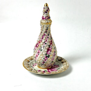Peut inclure: Une bouteille de parfum décorative avec sa soucoupe assortie. La bouteille est blanche avec un motif en spirale, ornée de motifs floraux roses et blancs et de bordures dorées. La soucoupe présente un motif floral assorti et des bordures dorées.