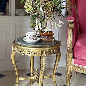 May include: A gold-painted, decorative side table with a circular, marble-effect top. A teacup and saucer with floral patterns are on the table, beside books and a vase of flowers. A pink chair is in the background.